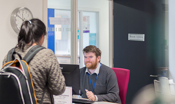 A ϲ staff member smiles up at a student who is in front of his desk