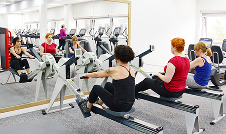 A row of women using rowing machines in front of a mirrored wall, ϲ