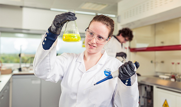 A ϲ student wearing PPE and holding up a beaker of yellow liquid