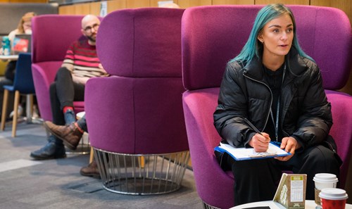 Student with vibrant blue hair sat on purple pod seat in ϲ Atrium